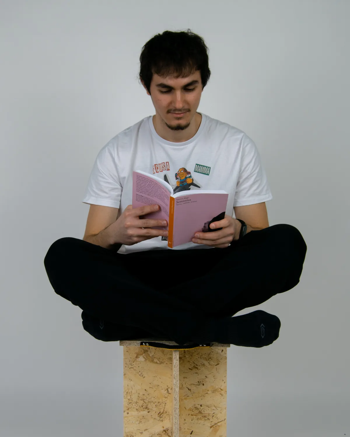 Riccardo Balletto seduto a gambe incrociate su uno sgabello di truciolare, legge un libro